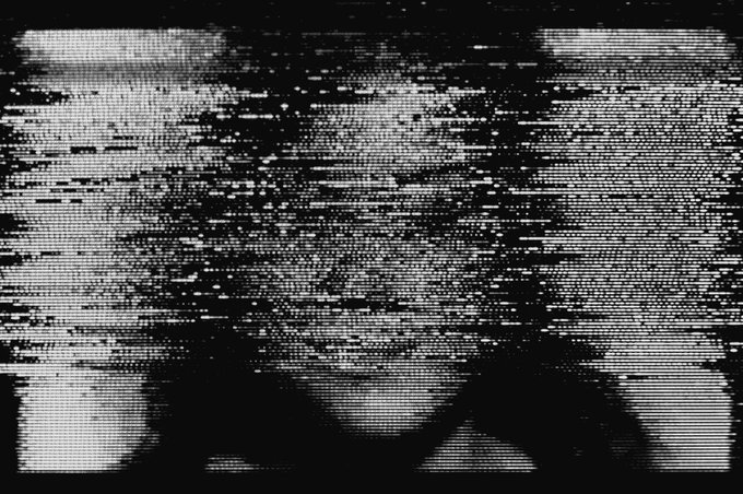 A black and white distorted/glitchy image with static noise patterns making the subject barely visible.