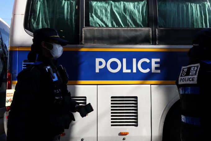 The image shows a police officer wearing full body armor, standing next to a large white bus with "POLICE" written across its side. Another person can be seen near the police officer, potentially a civilian or someone associated with the criminal organization being busted. The photo appears to capture an intense moment from the bust of the money laundering ring in Seoul, South Korea.