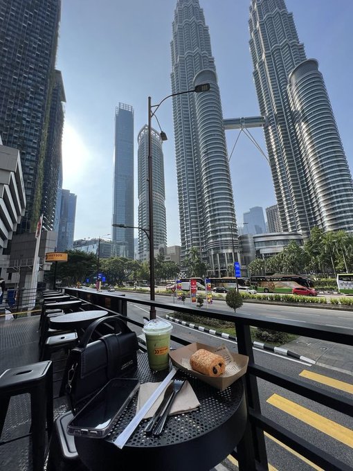 An outdoor cafe table with a matcha latte, a pastry, a smartphone, and a black bag sits in the foreground, with the iconic Petronas Twin Towers and surrounding Kuala Lumpur skyscrapers visible in the bright daytime background.
