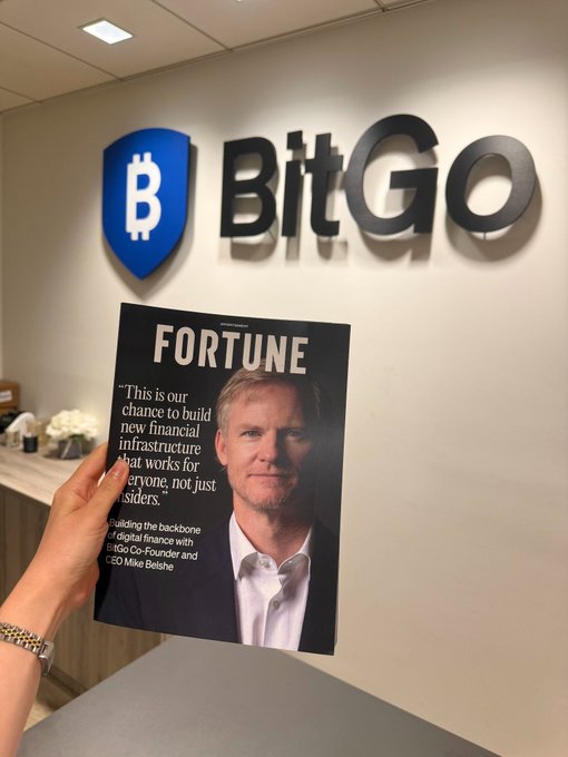 A hand holds a Fortune magazine featuring a quote from BitGo Co-Founder and CEO Mike Belshe about building financial infrastructure, photographed in front of the BitGo company logo on the wall—visually tying the magazine's feature to the "Fortune favors the BitGo" caption.