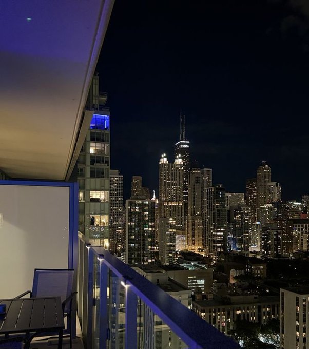 A nighttime view from a high-rise balcony overlooking a brightly lit city skyline, featuring a prominent twin-spired skyscraper and an outdoor chair on the balcony.