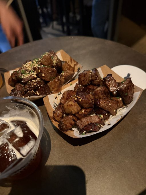 Two paper trays filled with charred wagyu beef bites sprinkled with sesame seeds and herbs sit on a dark table next to a plastic cup of iced beer, matching the relaxed Pumpfun event vibe described.