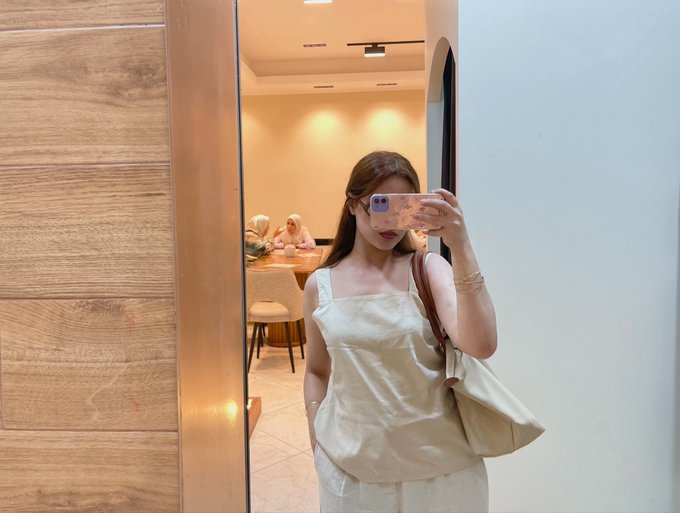 A mirror selfie of a woman taking a photo with a smartphone in a bright room. She is wearing a beige top and pants, carrying a cream-colored handbag, with other people visible seated at a table in the background reflection.