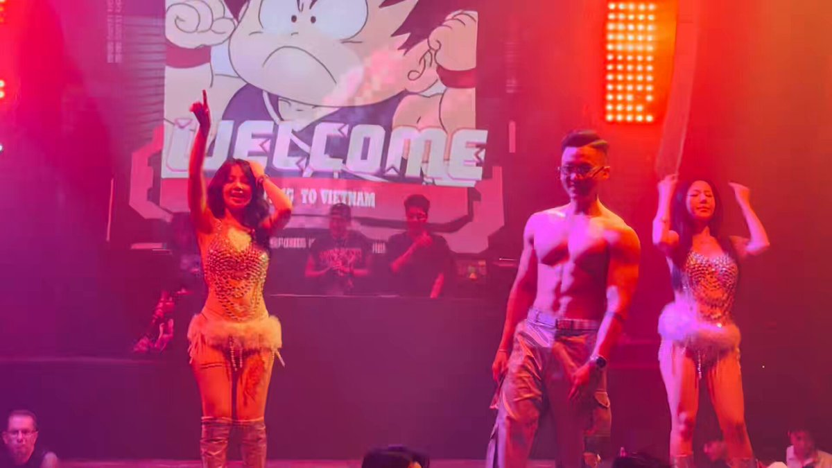 The image features three people standing on stage, dressed in costumes that include leotards for one person and a shirt and jeans for another. The third person appears to be a man wearing glasses. They are all smiling and appear to be enjoying themselves. In front of them is a large screen displaying the words "Welcome" with an image of a cartoon character. A banner reading "I'M SO happy today—my old friend came to hang out with me!" is also visible in the scene, suggesting that this gathering might be related to a performance or event where they are showcasing their talents and entertaining others.