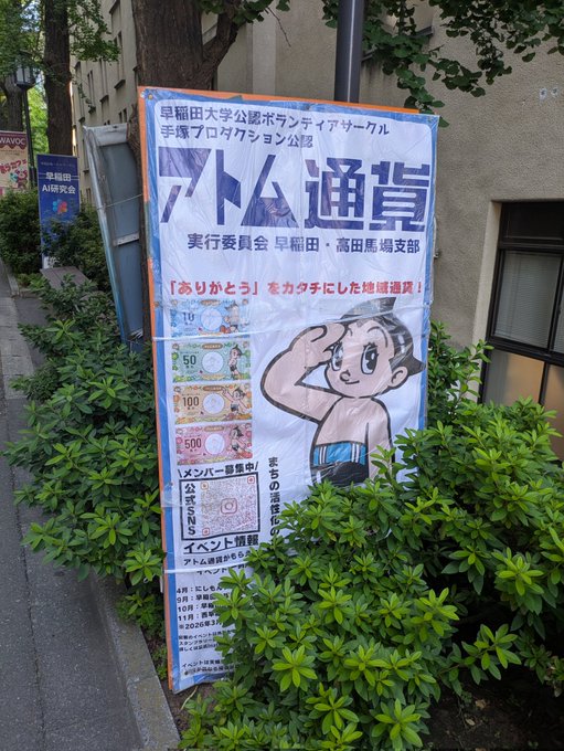 A promotional banner outside a Waseda University building advertises "アトム通貨" (Atom Currency) and "地域通貨" (local currency), featuring denominations of 10, 50, 100, and 500, along with a cartoon of the character Atom (Astro Boy). The banner promotes a Waseda University volunteer circle's local currency program, highlighting it as a real-world example of a community-based Japanese yen-pegged digital currency like JPYC.