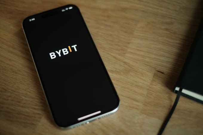 A smartphone rests on a wooden surface displaying the "BYBIT" logo on its screen, alongside a partially visible laptop with a connected cable.