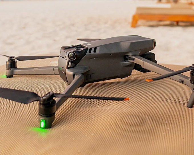 A dark gray DJI drone with extended propellers sitting on a textured mat at the beach, with green status lights illuminated and a wooden lounge chair blurred in the background.