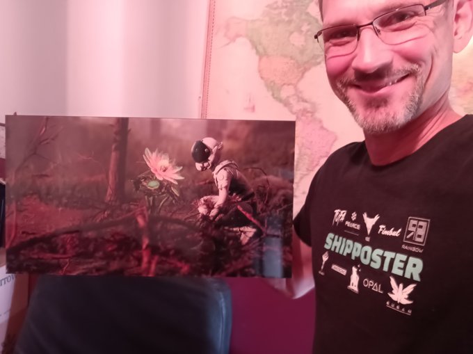A smiling man wearing glasses and a black 'SHIPPOSTER' t-shirt holds up an artwork depicting a small figure kneeling beside a glowing flower in a forest.