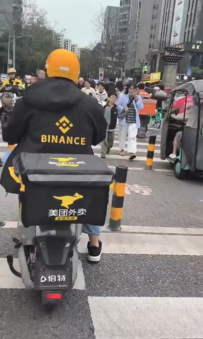 A delivery rider wearing a black “BINANCE” jacket and orange helmet rides a scooter with a “美团外卖” delivery box through a busy urban street.