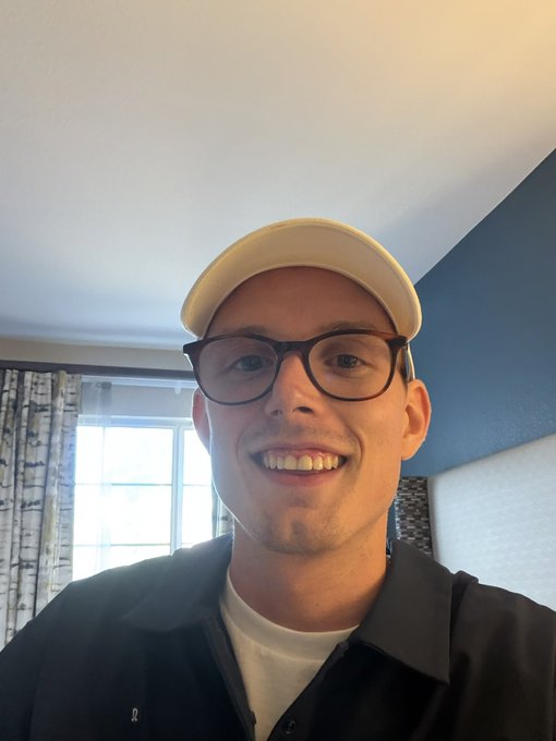 A selfie of a smiling man wearing black-rimmed glasses, a beige cap, and a dark jacket over a white shirt, with a window and curtains visible in the background.