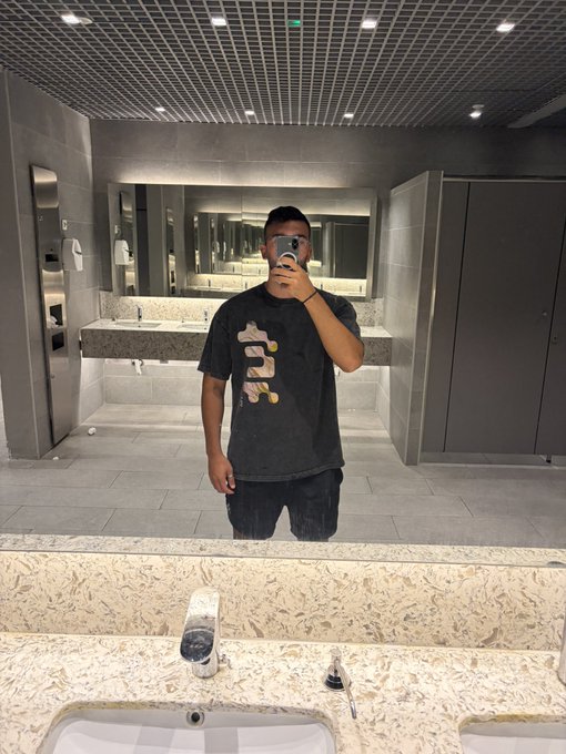 A mirror selfie of a person wearing a dark t-shirt with a colorful graphic, taken in a public restroom with sinks and stalls visible in the background.