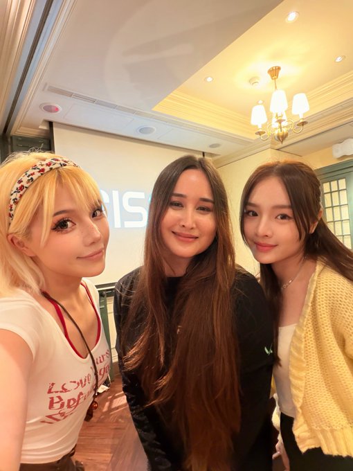 Three women pose together for a selfie indoors, with one wearing a floral headband and white t-shirt, another in a black top, and the third in a yellow cardigan, under an ornate chandelier with the partial text 'RIS' visible in the background.
