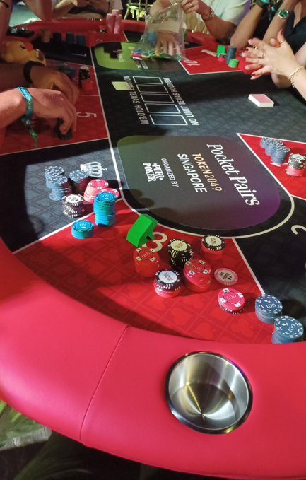 A red poker table is set up for a Texas Hold'em tournament, with stacks of colorful poker chips, player hands, and promotional text visible on the felt.