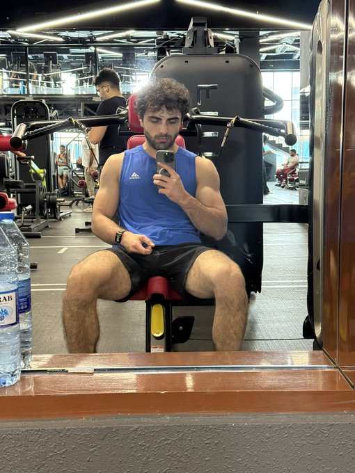 The image shows a man sitting on a weight machine at a gym, taking a selfie with his cell phone. The man appears to be wearing a blue shirt while using the exercise equipment. There are other people present in the background of the scene, indicating that this is likely a public fitness facility.