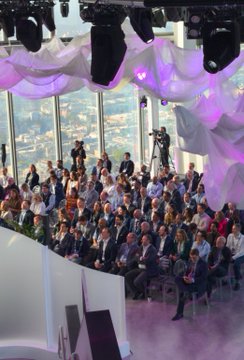 A formal event with a seated audience in business attire inside a modern venue with large windows and white draped decorations on the ceiling.
