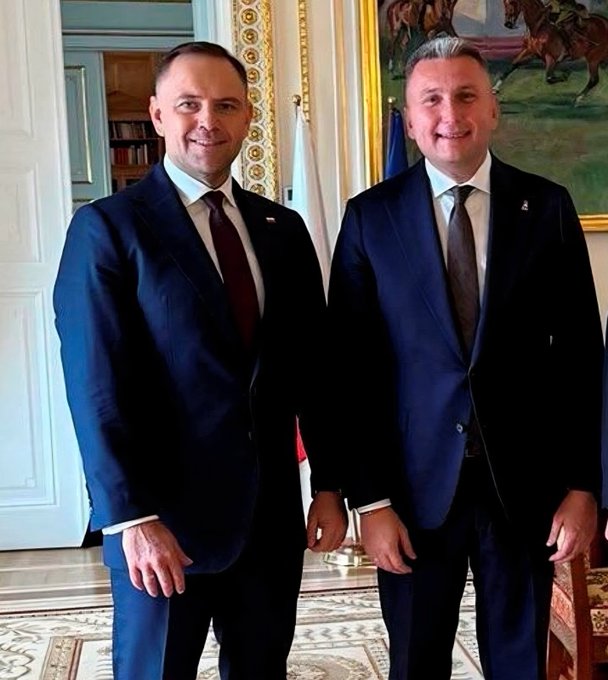 Two men in dark navy suits stand smiling side-by-side in an elegant, ornate room featuring classical architecture, gilded molding, and a large painting in the background. The image appears to capture a formal meeting, likely between a Zondacrypto representative and a government official, fitting the post's context about Polish cryptocurrency regulation discussions.