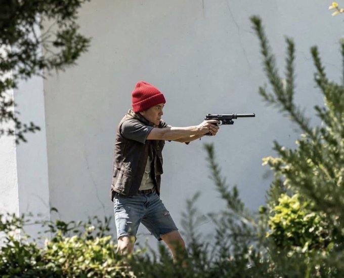A person wearing a red beanie, a brown vest, and denim shorts aims a black handgun outdoors, partially obscured by green foliage.