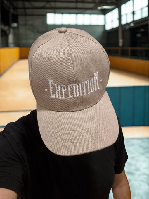 A close-up selfie of a person wearing a black shirt and a beige baseball cap with 'EXPEDITION.' embroidered in white, taken inside an indoor sports facility.
