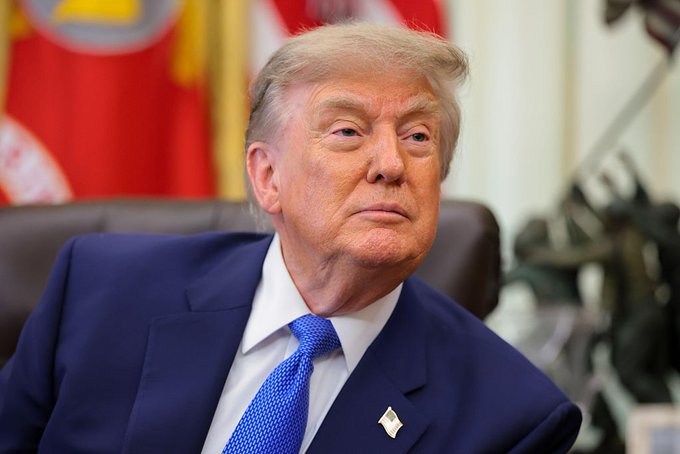 A close-up portrait of Donald Trump in a blue suit, sitting in a chair with flags in the background, likely related to his statements about cryptocurrency.