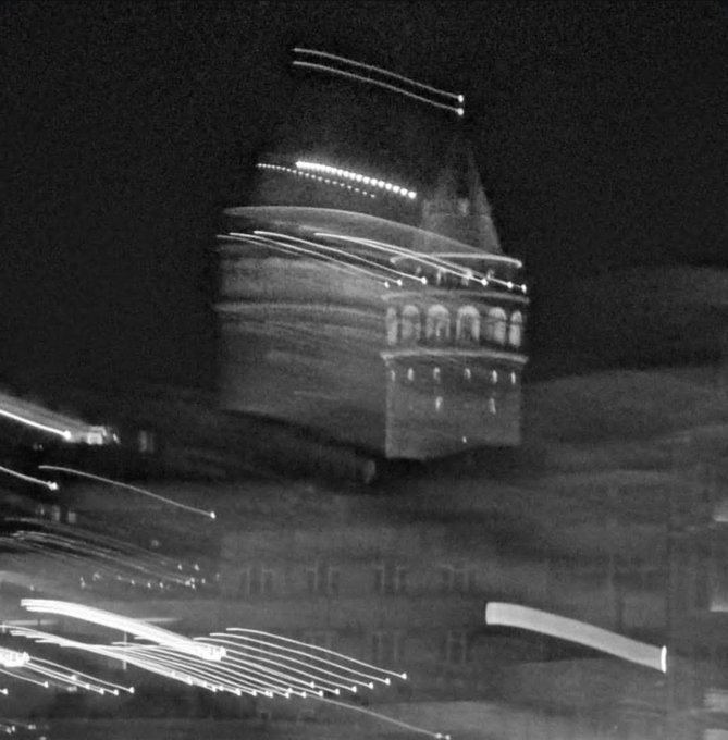 Black and white photograph with motion blur showing a large illuminated ship or building at night, with light trails streaking across the scene.