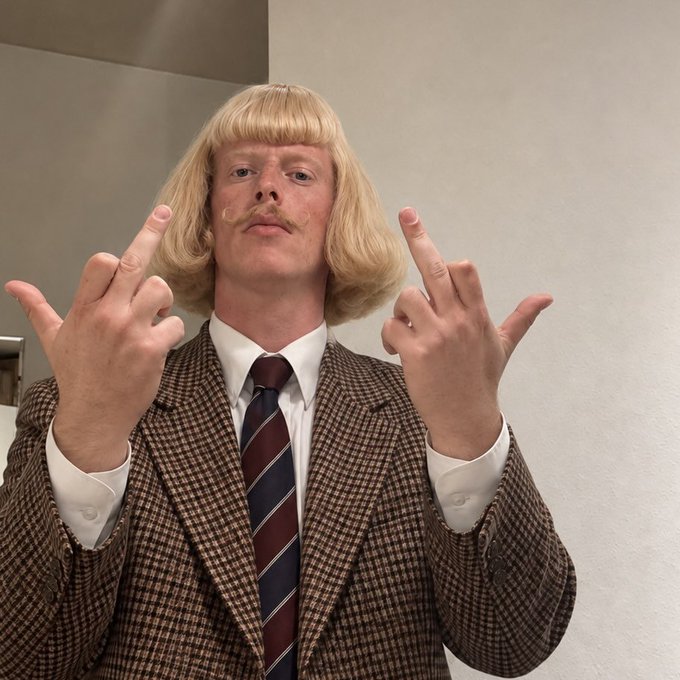 A person wearing a blonde bob wig, mustache, checkered blazer, and striped tie poses with both middle fingers raised.