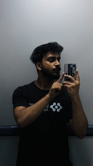 A young man with dark hair takes a mirror selfie with his phone while wearing a black t-shirt that has a small logo on it.