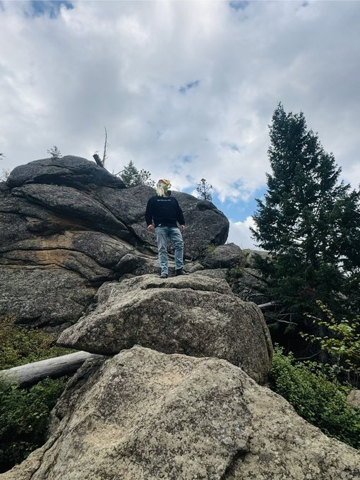 A low-angle photograph of a person wearing a dark top and jeans standing atop a large granite boulder formation, with pine trees and a cloudy sky visible in the background.