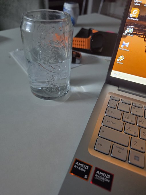 An empty glass of water sits beside an open laptop with "AMD Ryzen" and "AMD Radeon" stickers and a Thai-language desktop, while a "GM CT" post greets viewers with a casual workspace moment.