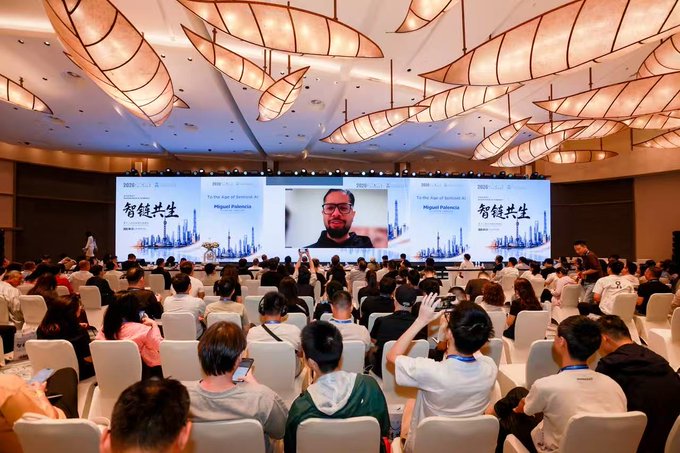 Large conference event featuring a speaker named Miguel Palencia displayed on a massive screen, with attendees seated in rows under an ornate ceiling decorated with leaf-shaped chandeliers, and Chinese text '智链共生' visible on the display.