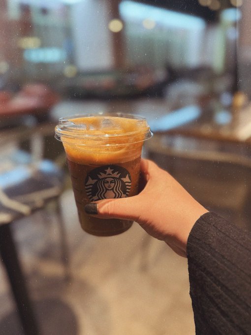 A hand with dark nail polish and a dark sleeve holds up a clear Starbucks plastic cup filled with an iced coffee drink inside a café.