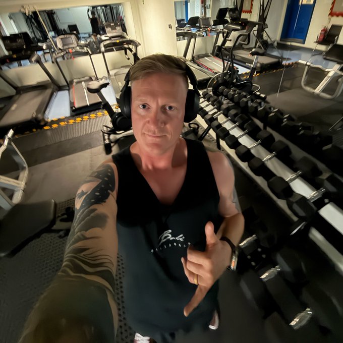 A tattooed man wearing headphones and a black tank top takes a selfie in a gym, with treadmills visible behind him and a long rack of dumbbells to his right.