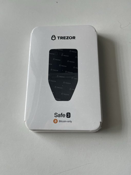 A white Trezor Safe 3 Bitcoin-only hardware wallet in its packaging, featuring a black shield-shaped design on the front.