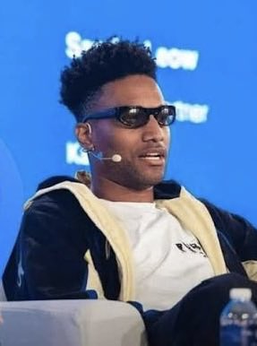 A young man with short curly hair, wearing dark sunglasses and a headset microphone, sits on stage against a blue backdrop, apparently speaking at a conference. The image accompanies breaking news that crypto influencer Ansem is predicting Bitcoin will reach $90,000 by the end of the week.
