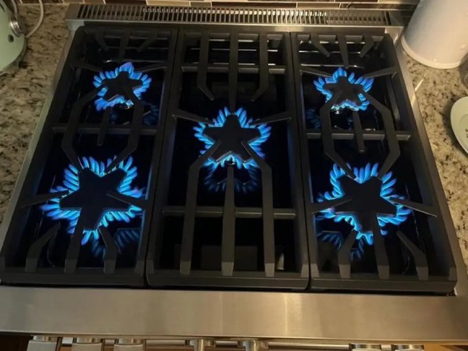 A six-burner gas stove with all burners lit, displaying vibrant blue flames forming star-shaped patterns on a black cast iron grate, set atop a stainless steel cooktop with granite countertops visible on either side.