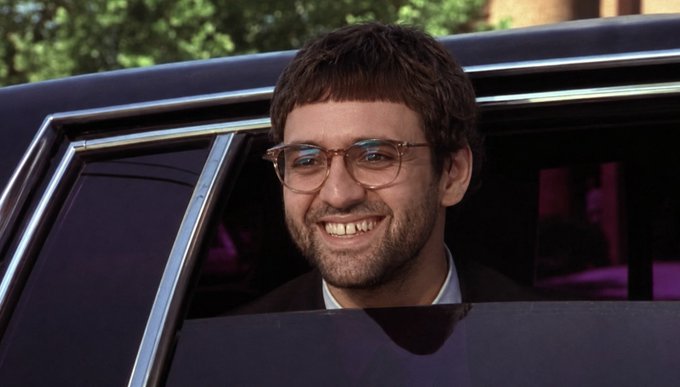 A man with short brown hair, a beard, and aviator glasses smiling while sitting in a car.