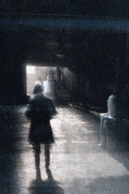 A grainy, dark image showing a child standing in a dimly lit room facing a doorway, with a chair visible to the right and other figures in the shadows.