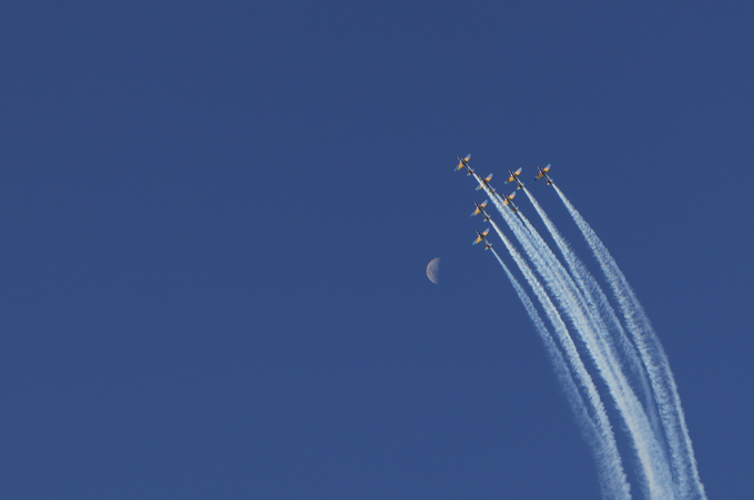 Six fighter jets fly in formation across a deep blue sky, leaving white contrails as they pass near a small crescent moon.