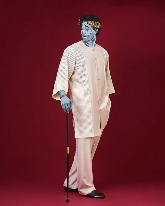 A fashion editorial photo shows a figure with blue-painted skin and a golden laurel wreath wearing a textured cream-colored outfit and holding a black cane against a dark red background.