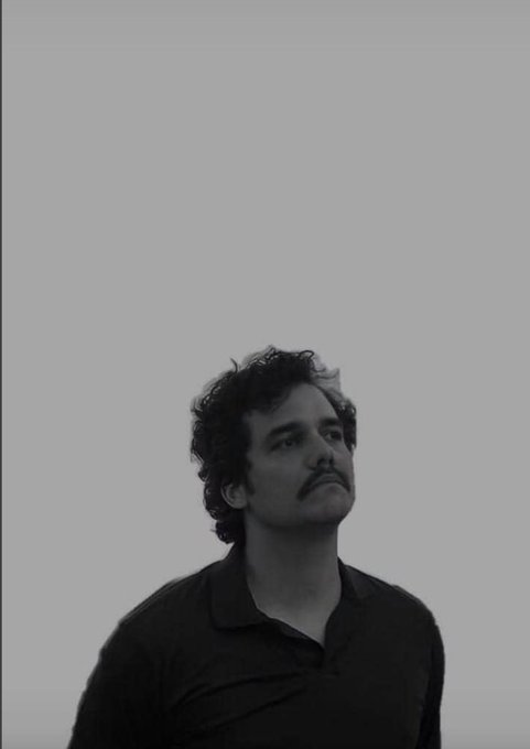 A black and white portrait of a man with curly hair and a mustache wearing a polo shirt, looking off to the side.