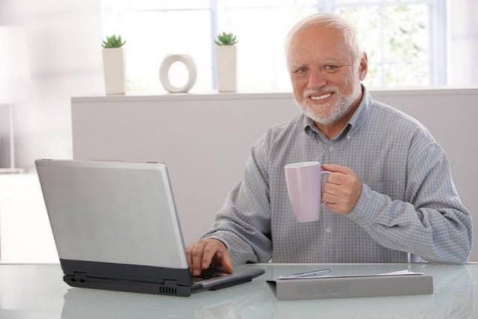 An older smiling man with a white beard using a laptop while holding a white mug in a bright kitchen.