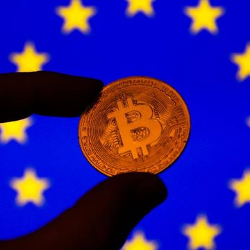 A close-up of fingers holding a physical Bitcoin coin against a backdrop of the European Union flag with its circle of yellow stars.
