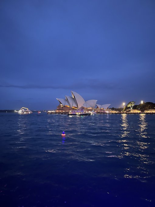 The Sydney Opera House, a white sailboat with a blue stripe, is reflected on the water's surface at dusk, with other boats and buildings visible in the background.