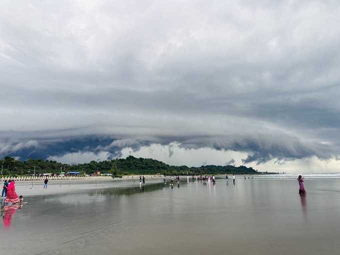 A dramatic coastal scene featuring dark, heavy storm clouds rolling in over a beach with people walking along the wet sand and shallow water reflecting the sky.