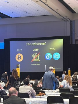 The image shows a group of people sitting at tables with chairs arranged around them. The room has a large screen displaying information about Bitcoin and its impact on financial freedom, as well as an exit sign indicating that the event is over. A man wearing a suit can be seen in the foreground, while other attendees are seated throughout the scene.