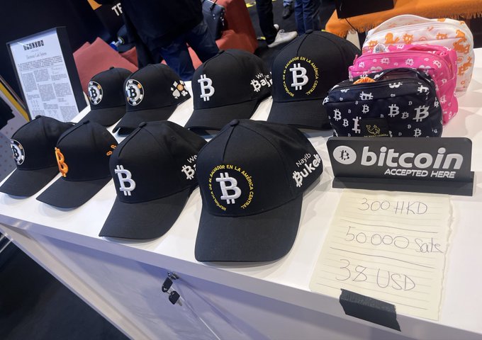 A display table featuring black baseball caps with the Bitcoin 'B' logo, alongside a sign reading 'Bitcoin ACCEPTED HERE' and a handwritten price list showing '300 HKD', '50 000 Sats', and '38 USD'.