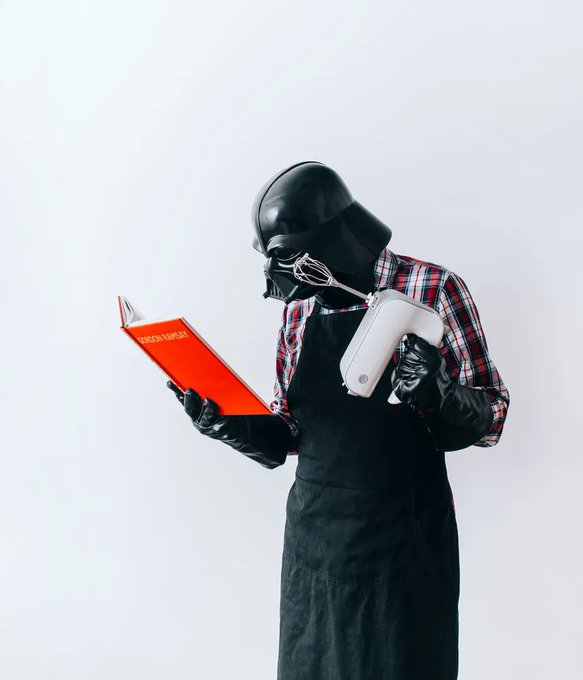 A person dressed in a Darth Vader mask, plaid shirt, and apron holds an open red cookbook while operating a white hand mixer, channeling a "dark side" cooking session in classic "be right back" fashion.