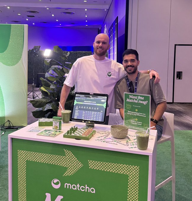 The image shows two men standing behind a booth at an event, with a green sign that says "matcha" written on it. The booth appears to be promoting matcha products or services related to the event. One of the men has his arm around another man's shoulder in a friendly manner, suggesting they are engaging with each other and possibly discussing the booth offerings.