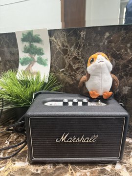 A black Marshall speaker sits on a marble surface next to a small plush hamster toy, with a bonsai tree and bonsai painting visible in the background.