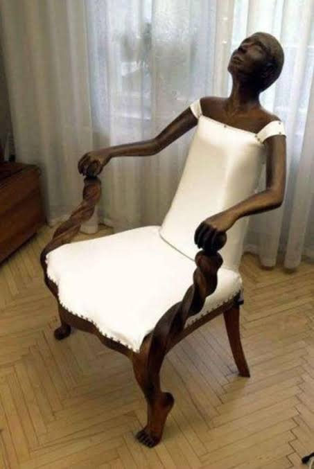 A dark, intricately carved wooden chair with white upholstery that incorporates a sculpted figure of an emaciated person leaning back with their head tilted upward.