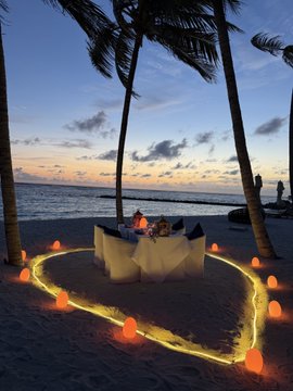 The image shows an outdoor dining area with a table set up for two, surrounded by palm trees. The table has white plates and napkins, creating a romantic atmosphere. There are orange lights strung across the sand that illuminate the scene, adding warmth and ambiance. A couple is seated at the table, enjoying their time together in this intimate setting. In the background, there's water visible, suggesting an ocean view or beach location. The post features information about converting spending on stablecoins to US dollars using a specific app called KAST, which offers cashback rewards for users who spend money through the app.